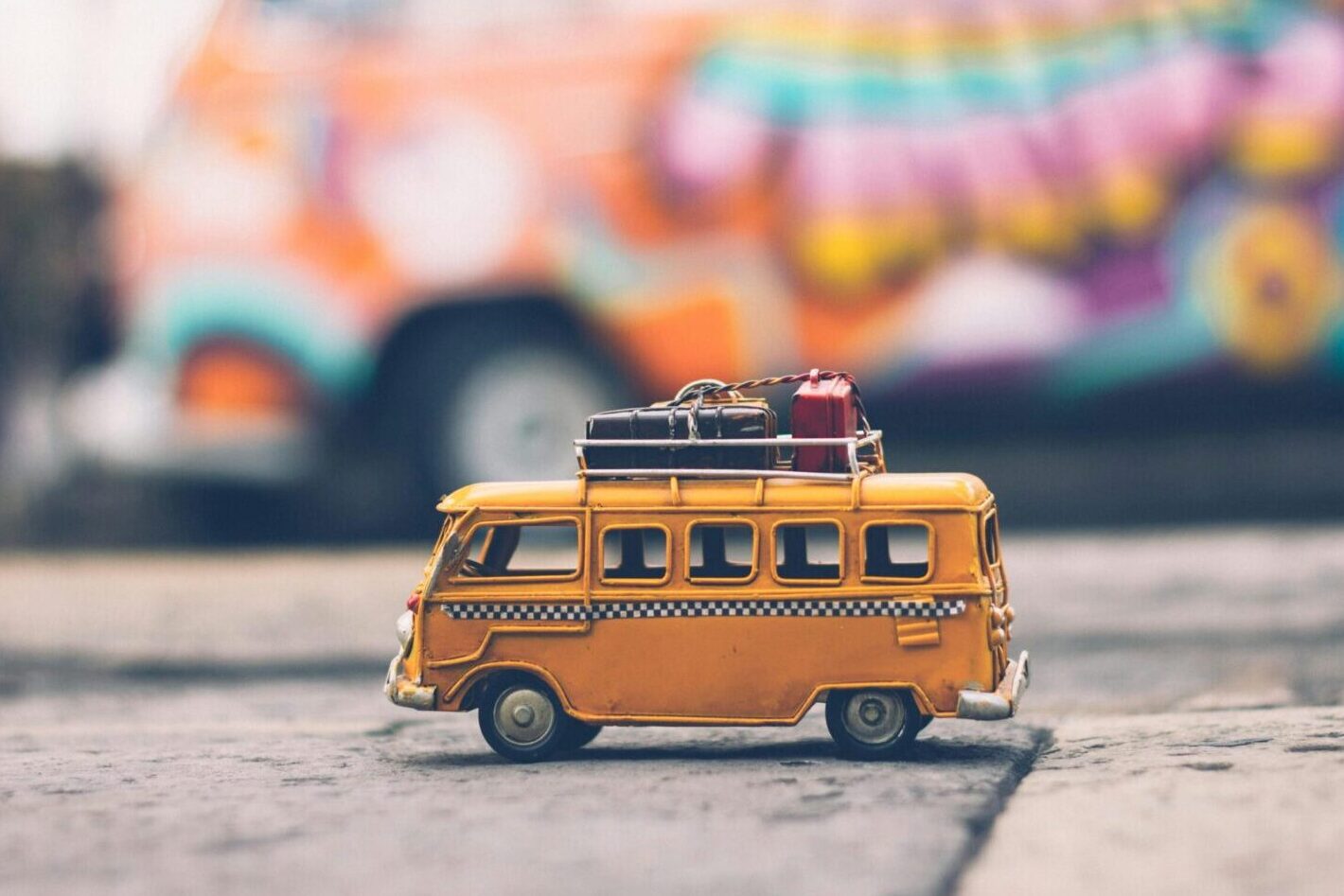 Close-up of a toy Volkswagen van with luggage, set against a colorful street backdrop, evoking summer travel vibes.